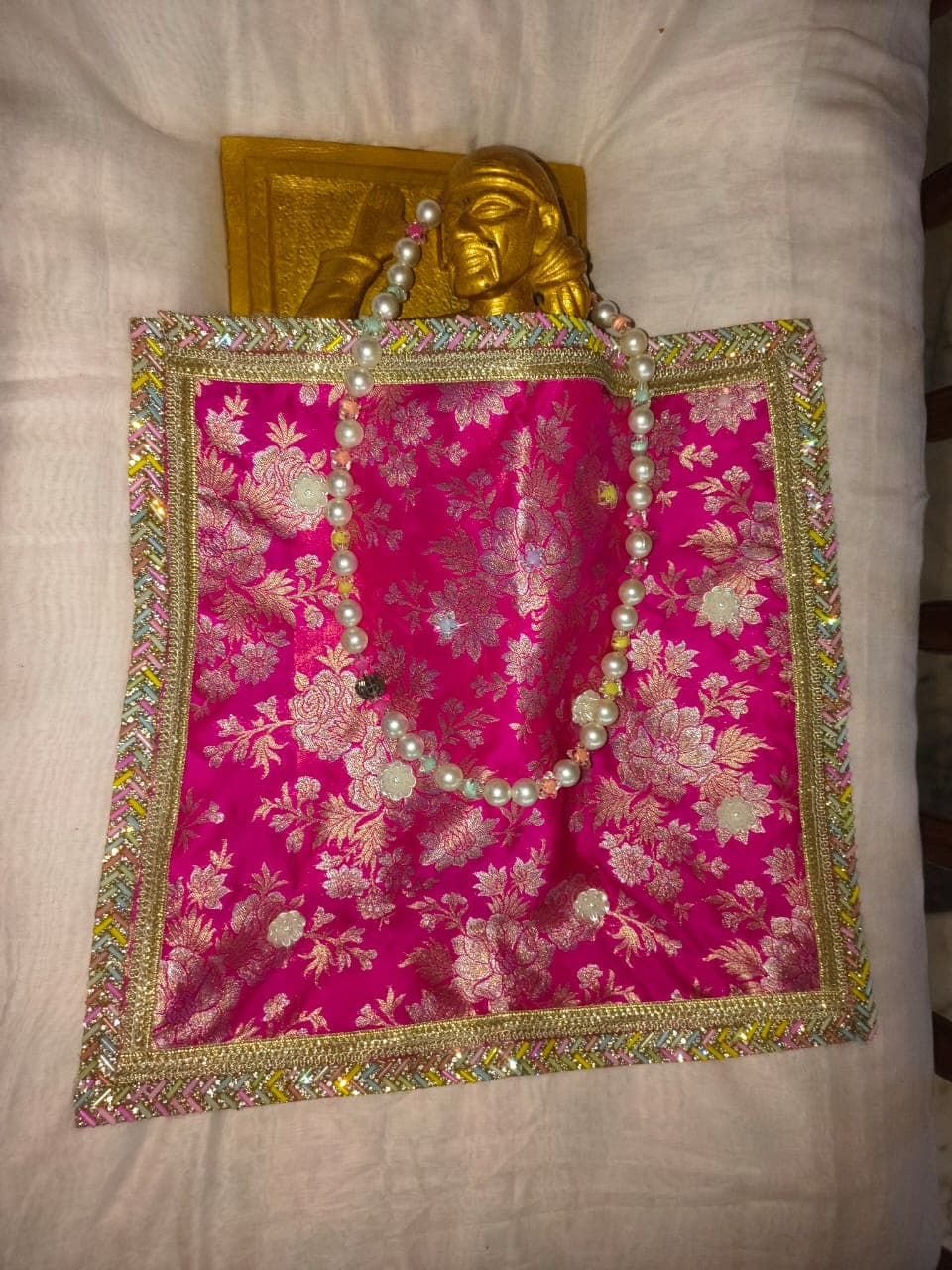 A pink floral fabric and pearl garland laid out beside a small gold murti head, prepared ahead of dressing the murti