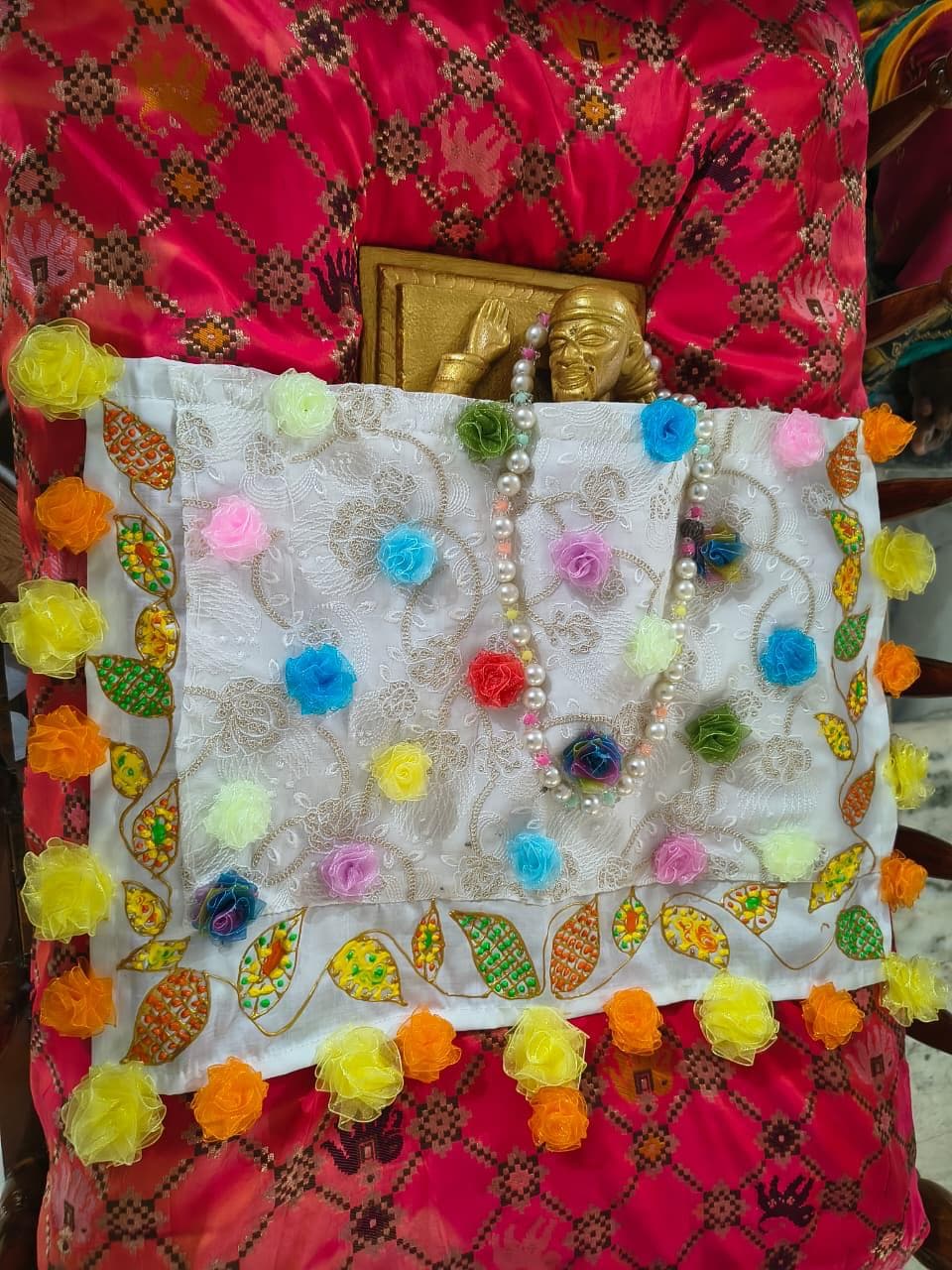 A white lace cloth decorated with colorful fabric roses and a pearl garland, laid over a red patterned fabric beside a small gold murti head, prepared ahead of dressing the murti