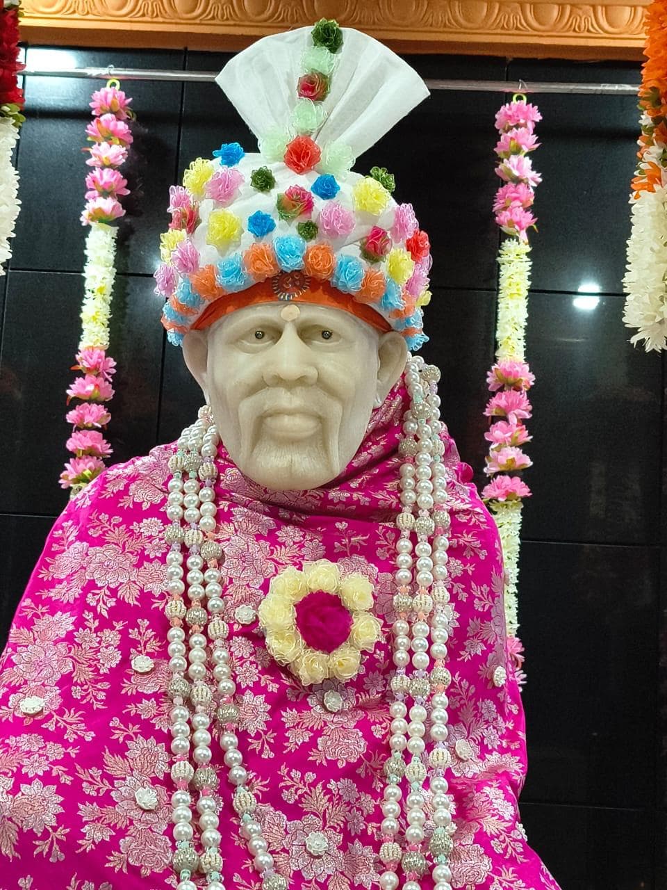 Close view of the murti's face and an ornate multicolor floral crown, dressed in a magenta floral-brocade shawl with layered pearl garlands and a red and yellow flower brooch