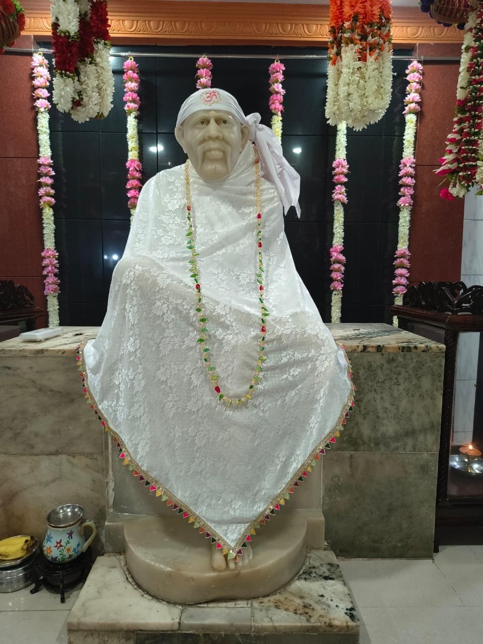 Close view of the murti's face and a white lace shawl marked with "ॐ" on the head-cloth, wearing a multicolor beaded garland