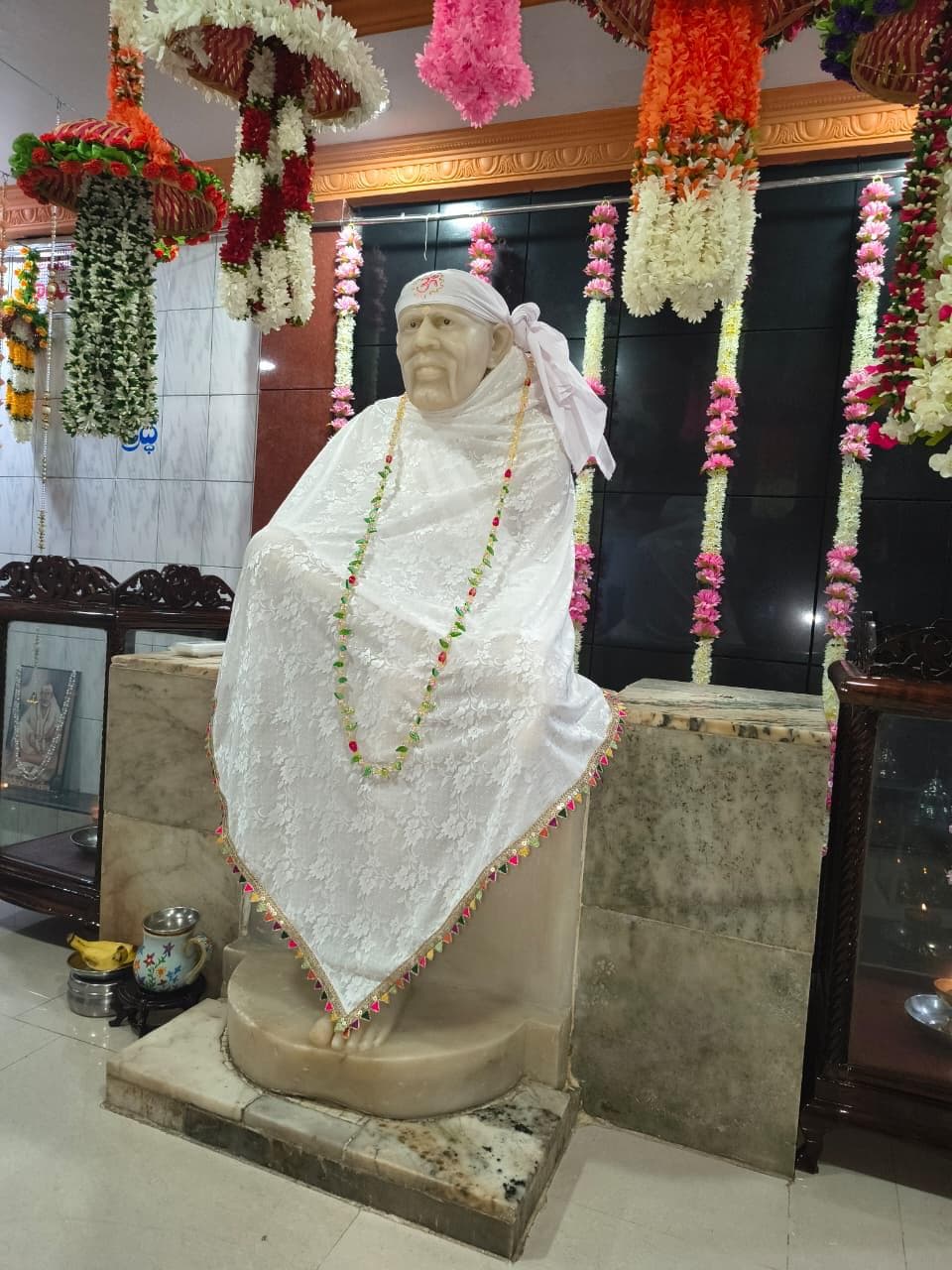 Another view of the murti in the white lace shawl and beaded garland, seen at an angle with hanging floral garlands behind