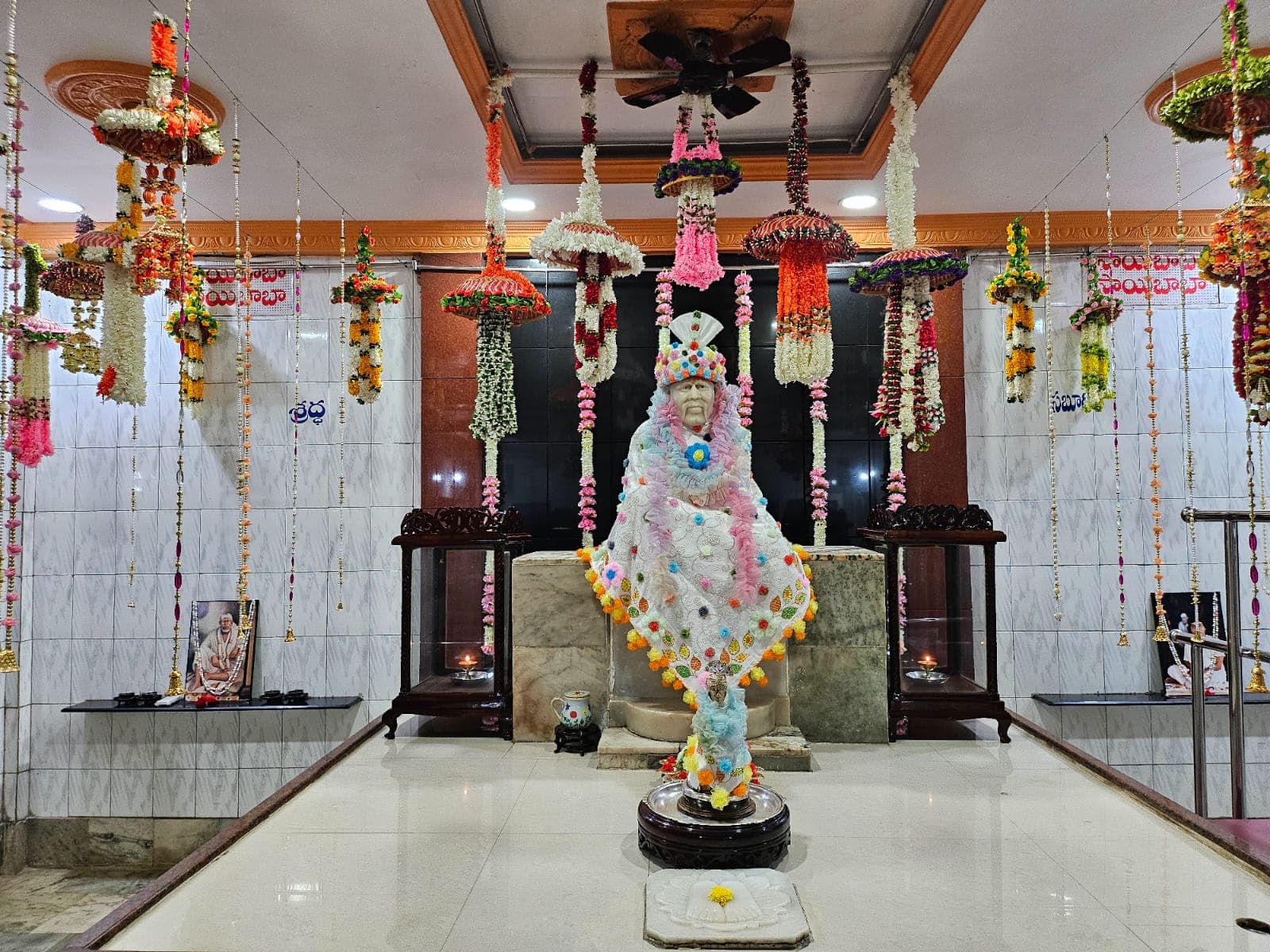 Wide view of the murti dressed in a multicolor floral poncho and beaded crown, hanging garlands throughout the sanctum