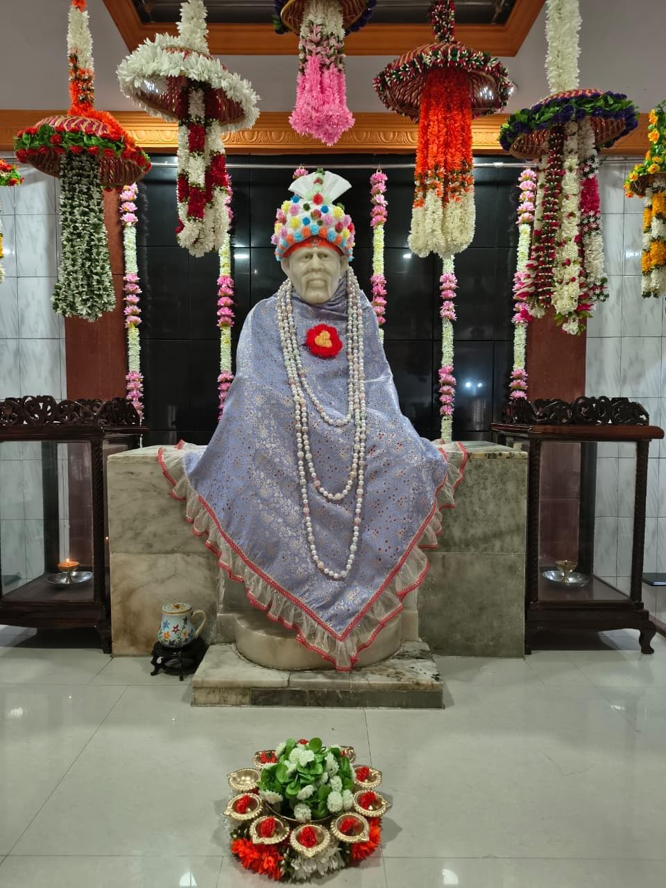 Wide view of the murti dressed in a lavender floral-brocade shawl with a pink ruffled trim, layered pearl garlands and a red flower brooch, hanging floral umbrellas above and a flower offering at the base