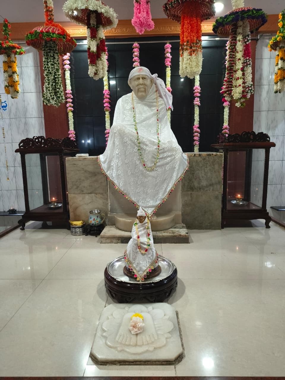 Wide view of the sanctum for Dasara, the murti dressed in a white lace shawl with an "ॐ" mark on the head-cloth and a multicolor beaded garland, the small processional murti dressed to match at the base, hanging floral umbrellas above