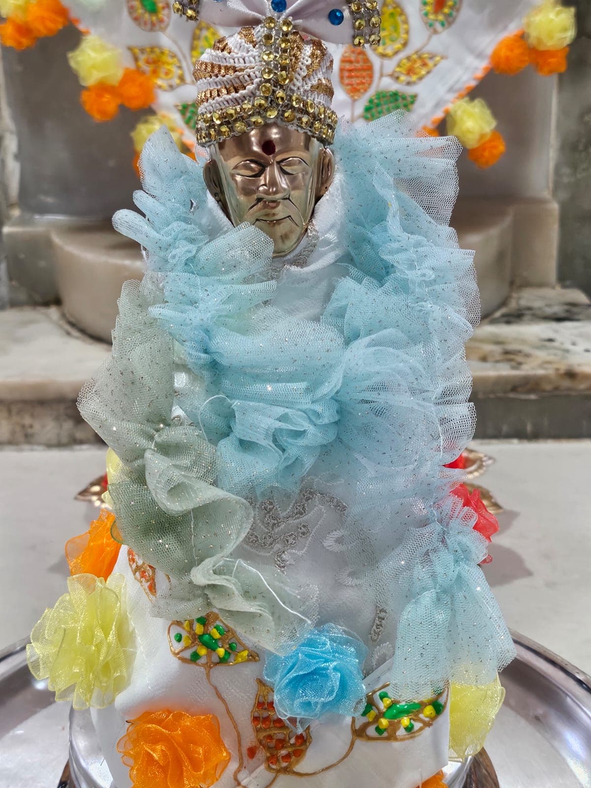 Extreme close-up of the temple's small metal murti, dressed in a pale blue ruffled fabric