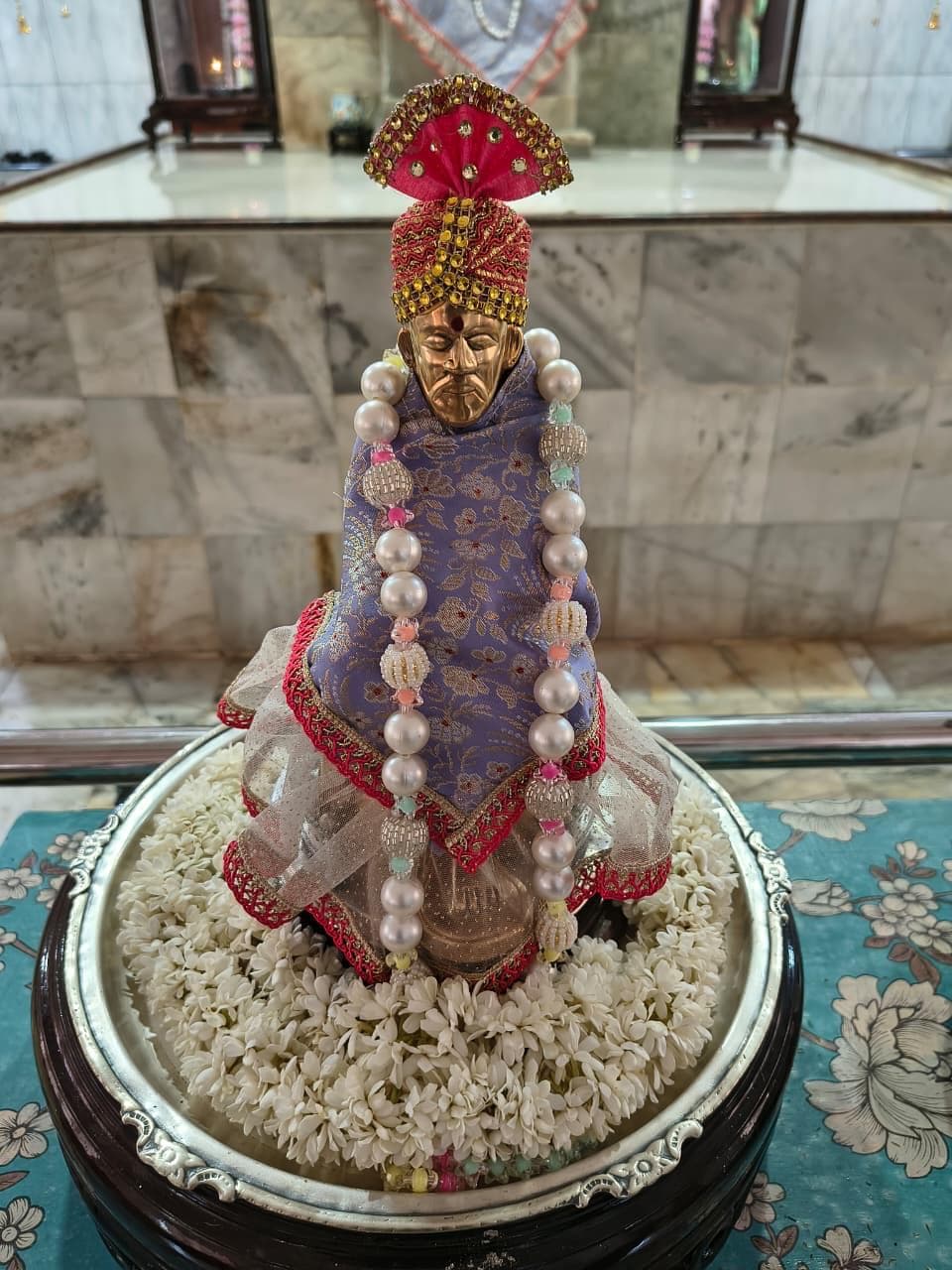 Close view of the temple's small processional murti (Utsava Vigraha), dressed in a lavender floral-patterned cloth with a red trim and pearl garland, set among white jasmine flowers