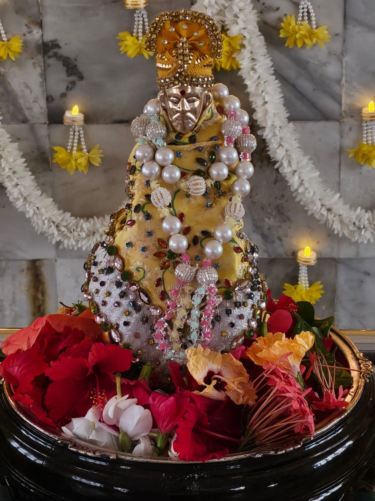 The temple's small golden murti dressed in a yellow jeweled shawl with layered pearl garlands, set in a tray of fresh flowers