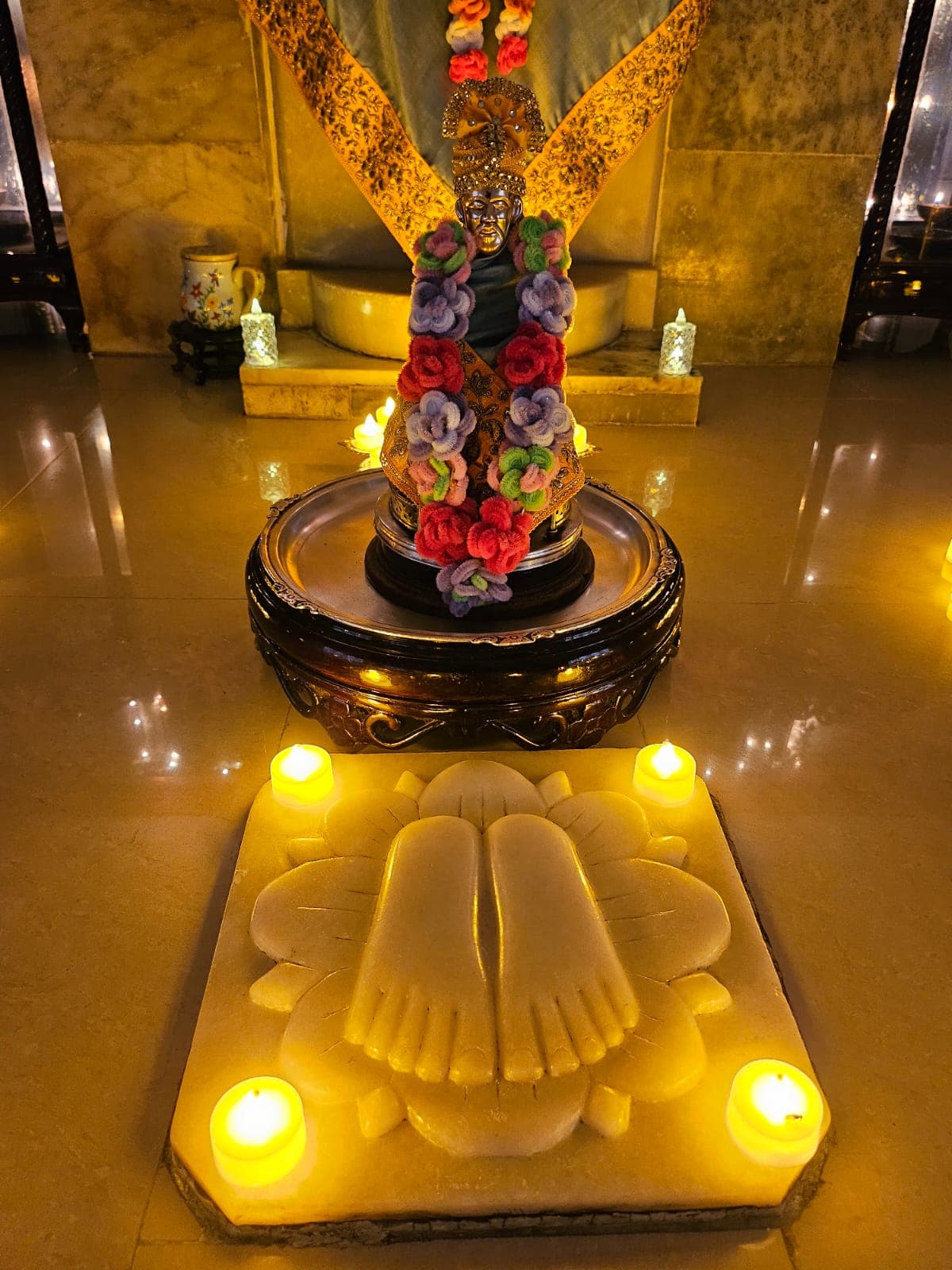 Close view of the temple's small dark metal processional murti, wearing an ornate gold crown and a teal and gold shawl, decorated with colorful crocheted flower garlands, set before the carved marble footprints with tealight candles