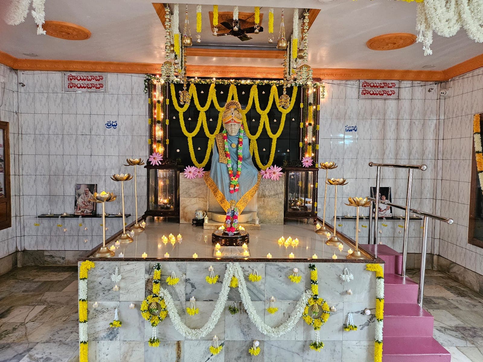 Sai Baba murti dressed in a blue shawl with a gold peacock-crest turban and a colorful flower garland