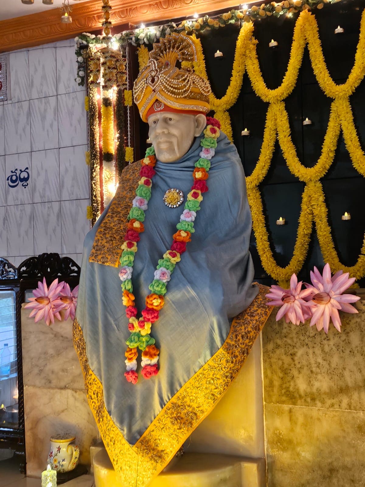 The murti in a blue shawl with a gold peacock-crest turban and a colorful flower garland, seen at an angle with the Shraddha signage visible on the wall and pink lotus flowers at the base