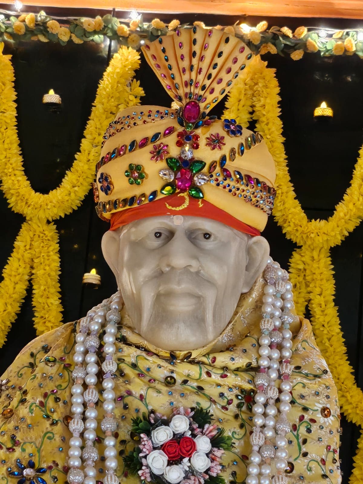 Close view of the murti's face, ornate gold turban, and yellow jeweled shawl, against a marigold backdrop