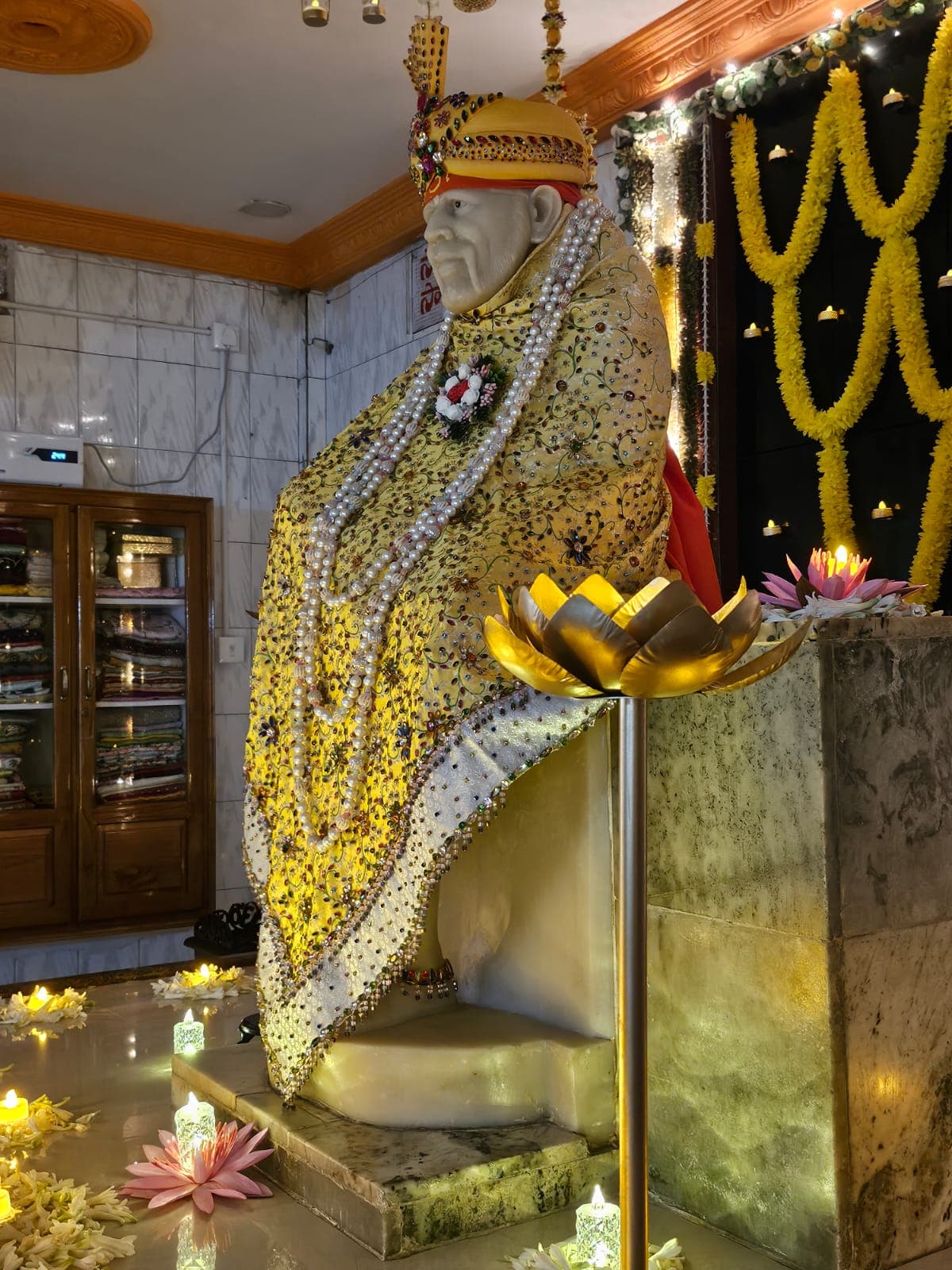 Sai Baba murti dressed in a yellow jeweled shawl with pearl garlands, against a yellow marigold backdrop
