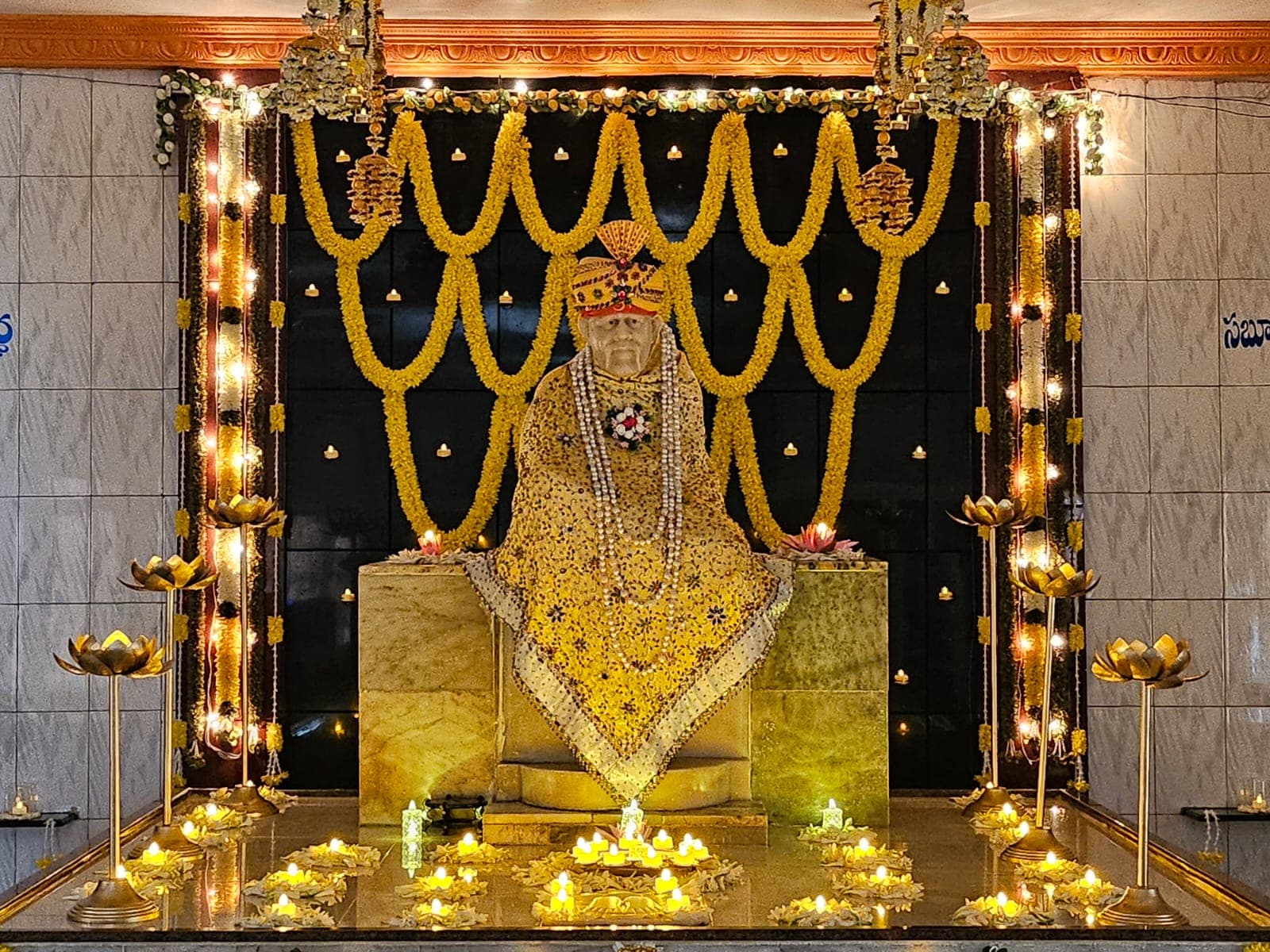 Wide view of the sanctum decorated for Guru Purnima, with the murti dressed in a yellow jeweled shawl beneath hanging marigold garlands, lit brass lotus lamps and diyas in front, and the Saburi signage visible on the wall