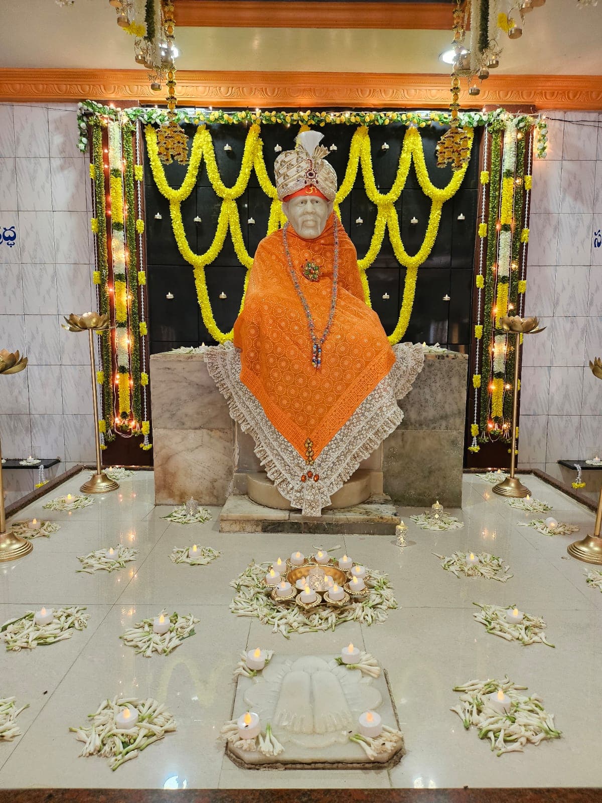 Sai Baba murti dressed in an orange shawl with a white lace border, garlanded with a beaded necklace