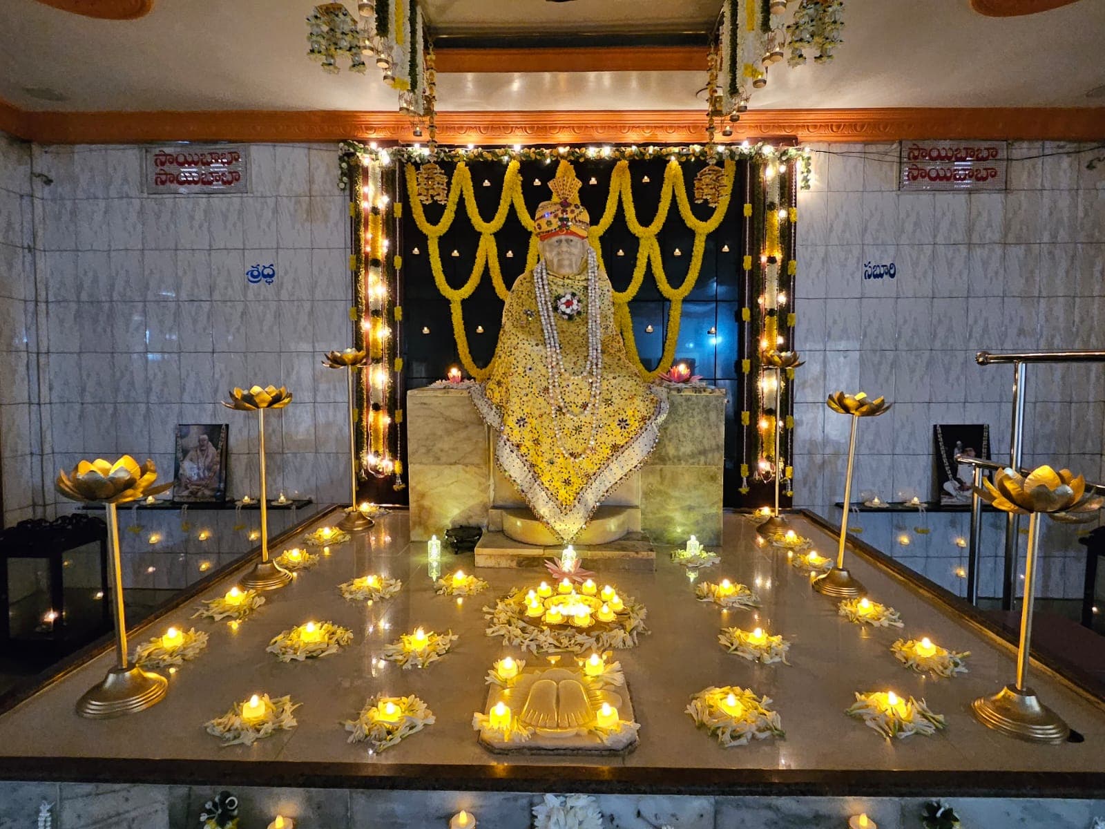 Wide view of the sanctum for Guru Purnima, the murti in a yellow jeweled shawl framed by hanging marigold garlands, with rows of tealight candles and flower petals arranged around Baba's carved footprints on the platform below, Shraddha and Saburi signage on the side walls