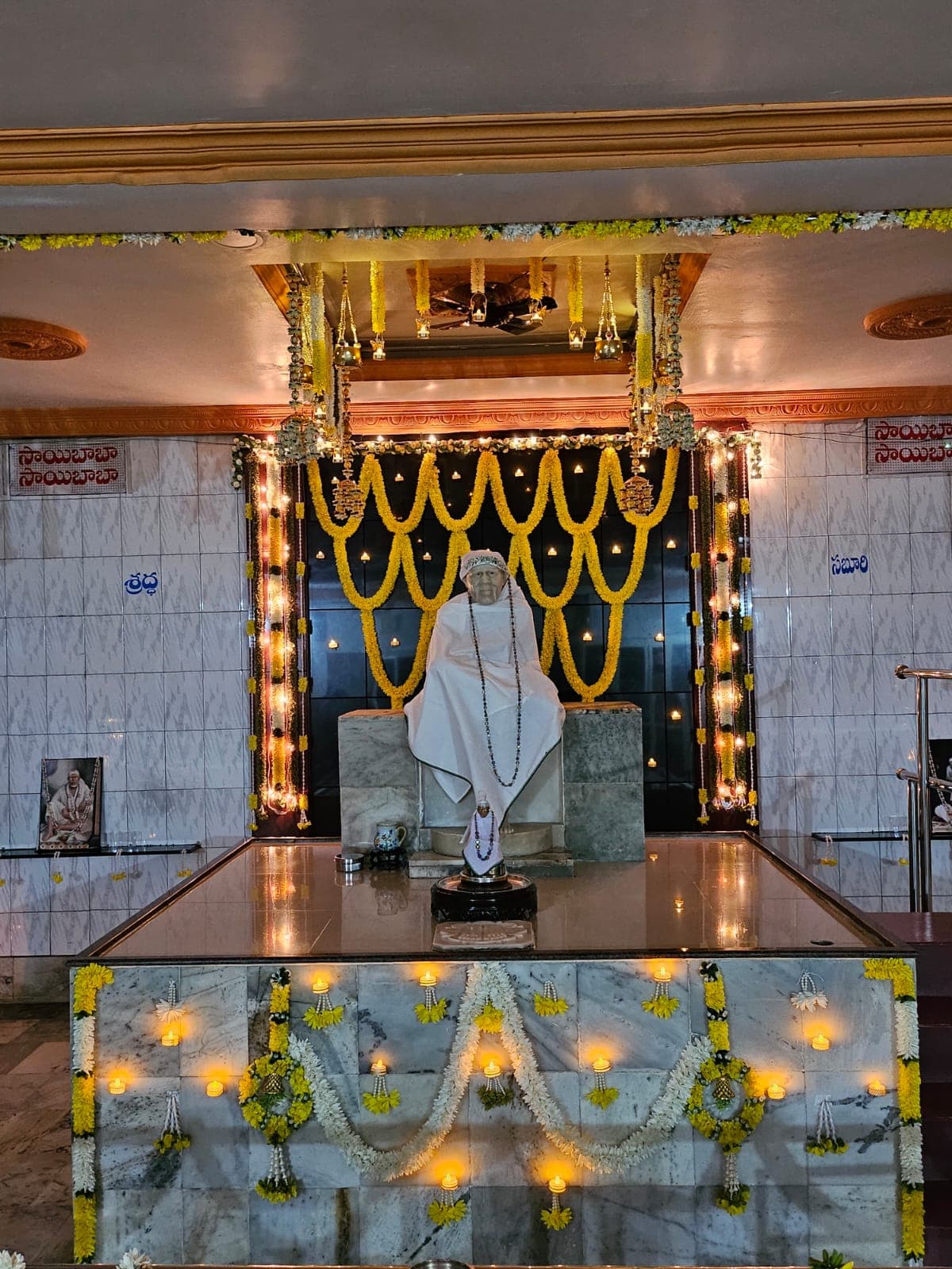 Sai Baba murti dressed in a plain white shawl, with the Shraddha and Saburi panels visible on either side