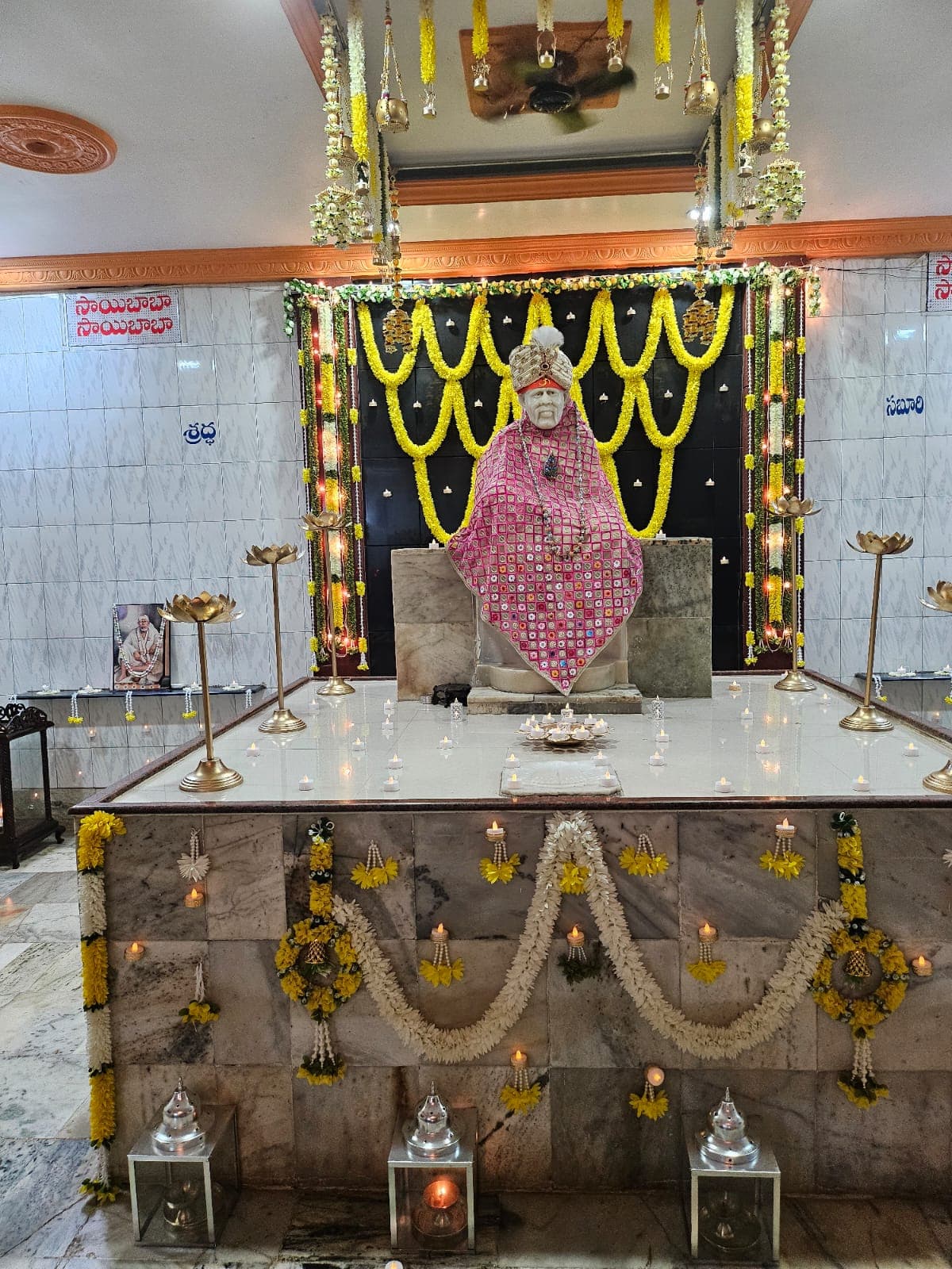 Sai Baba murti dressed in a pink jeweled shawl, with the Shraddha and Saburi panels visible on either side