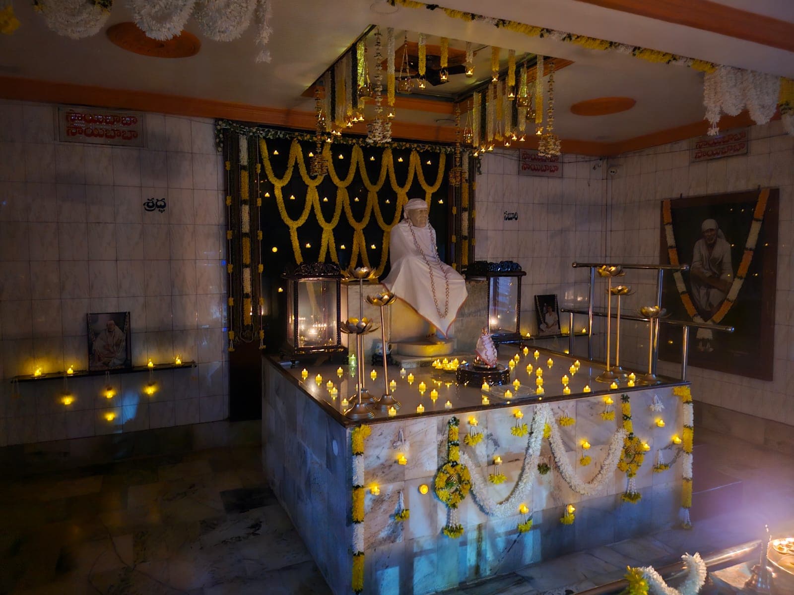 Angled wide view of the sanctum for Guru Purnima, the murti dressed in a plain white shawl beneath hanging marigold garlands, the Shraddha and Sabari signage visible on the side walls, the small processional murti also dressed in white at the base, tealight candles throughout