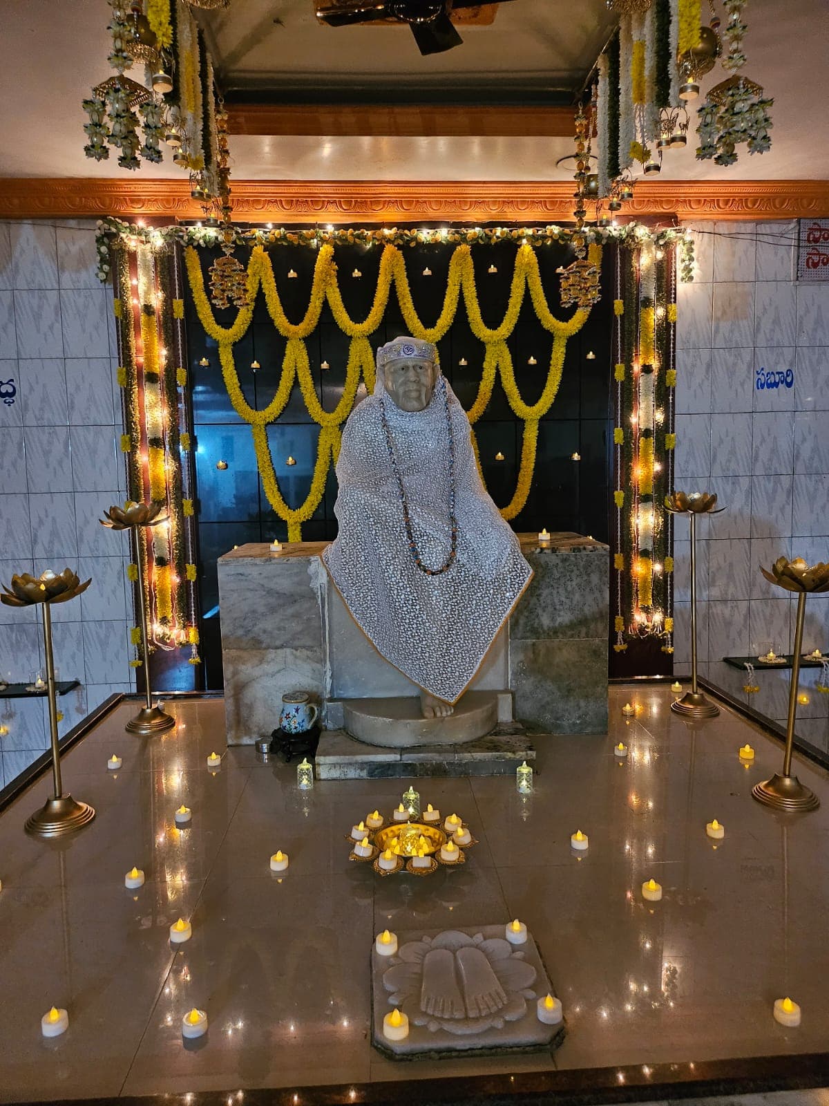 Wide view of the sanctum for Guru Purnima, the murti dressed in a textured silver-white shawl and cap marked with "ॐ", wearing a beaded garland, beneath hanging marigold garlands with brass lotus lamps and tealight candles arranged across the platform