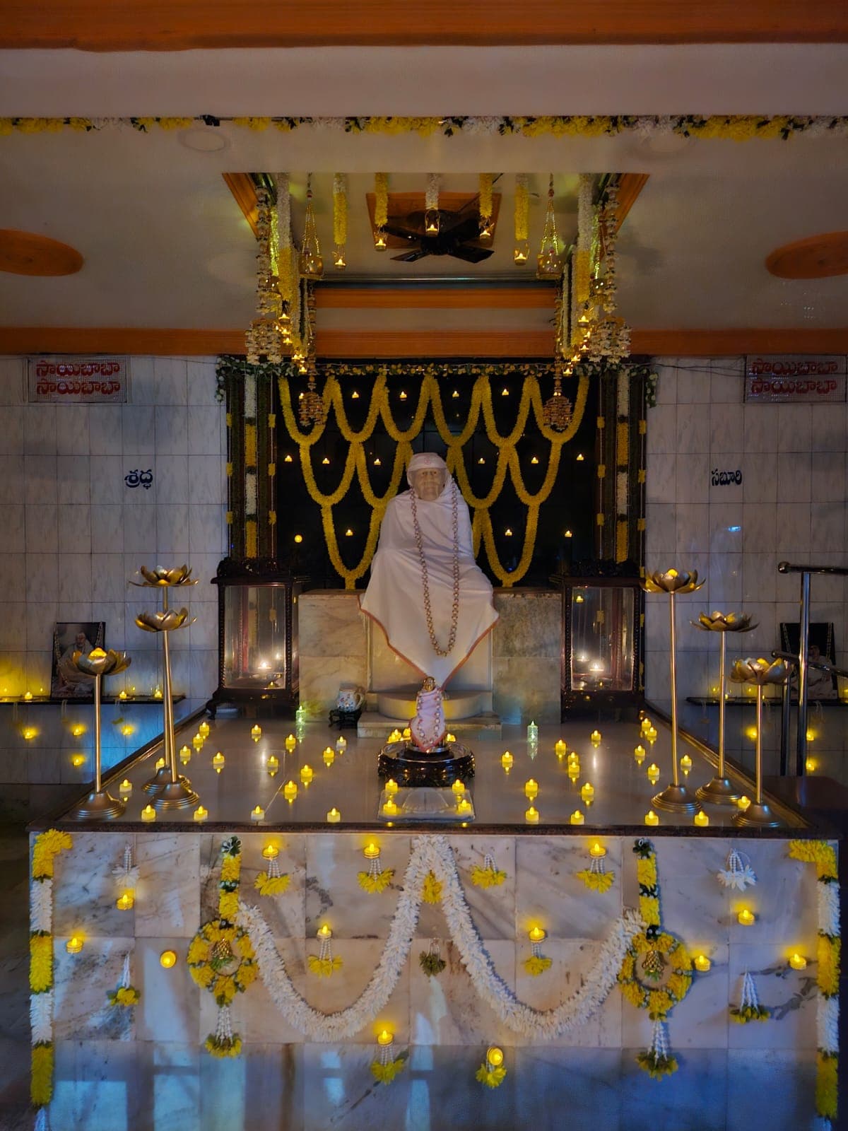 Wide view of the sanctum for Guru Purnima, the murti in a plain white shawl and pearl garland beneath hanging marigold garlands, with a small pink-clothed idol at the base and tealight candles arranged across the platform