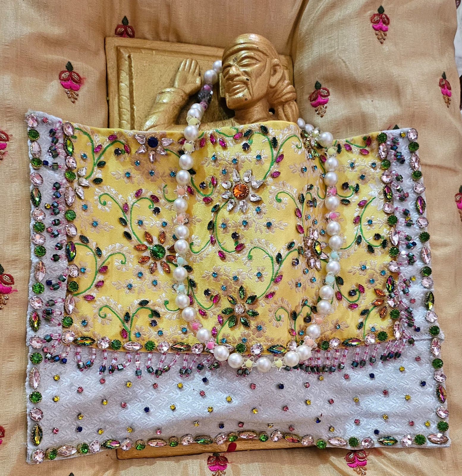 A jeweled yellow and white embroidered shawl with a pearl garland laid out in front of a small carved wooden Sai Baba figure and feet, prepared ahead of dressing the murti for Guru Purnima