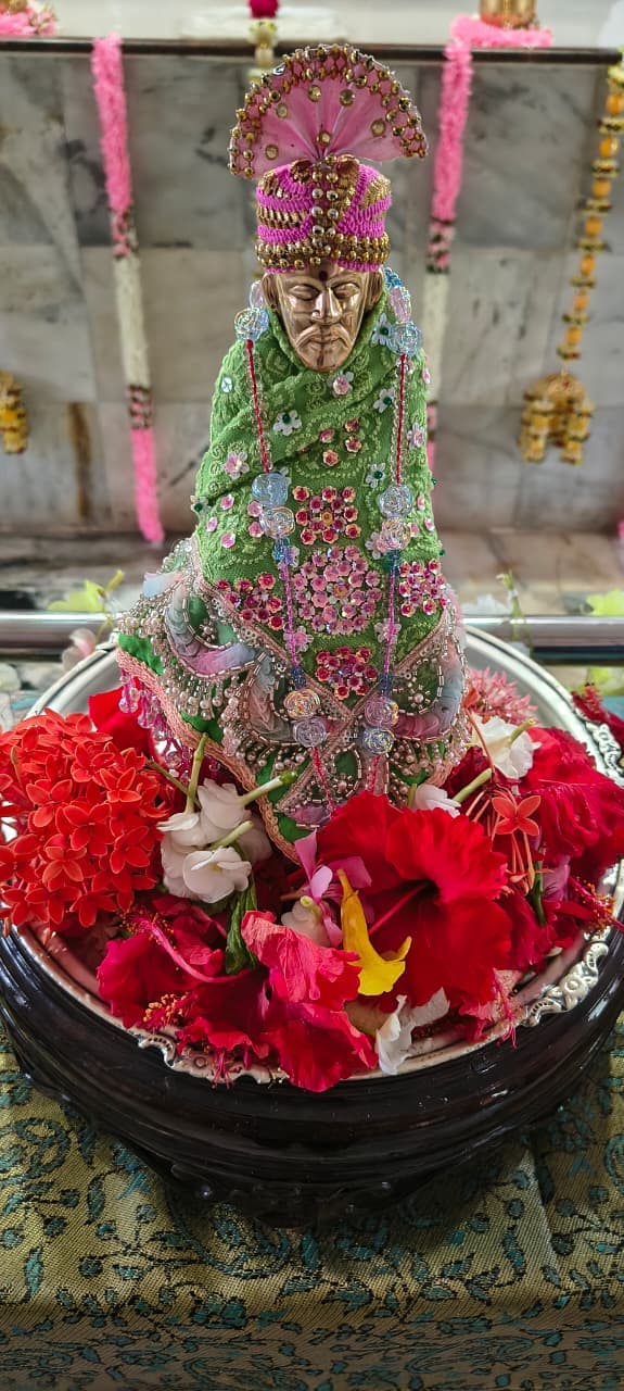 The temple's small murti dressed in a green jeweled shawl with a pink beaded turban, set in a tray of red, white, and yellow flowers