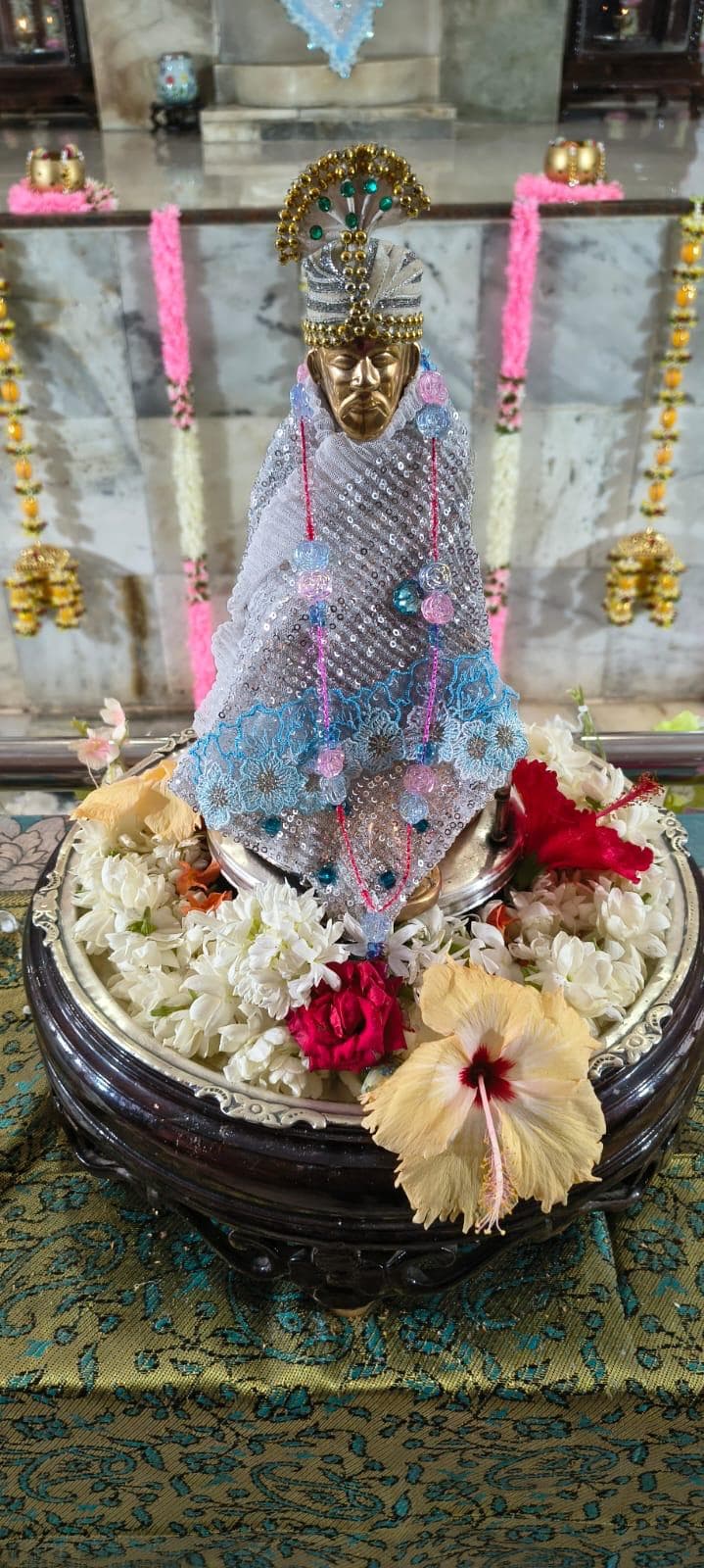 Close view of the temple's small golden-faced processional murti (Utsava Vigraha), dressed in a silver sequined cloth with a light blue lace trim and pink beaded garlands, set in a bed of white, red, and yellow flowers