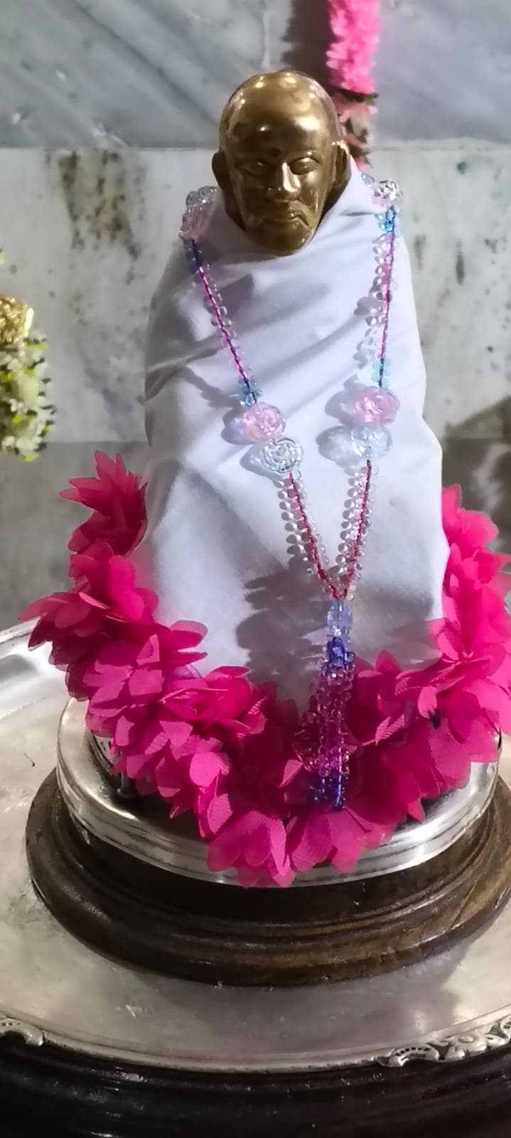 Close view of the temple's small golden-faced processional murti, dressed in a plain white cloth with a hot pink ruffled trim and beaded garland, set in a wreath of pink flowers