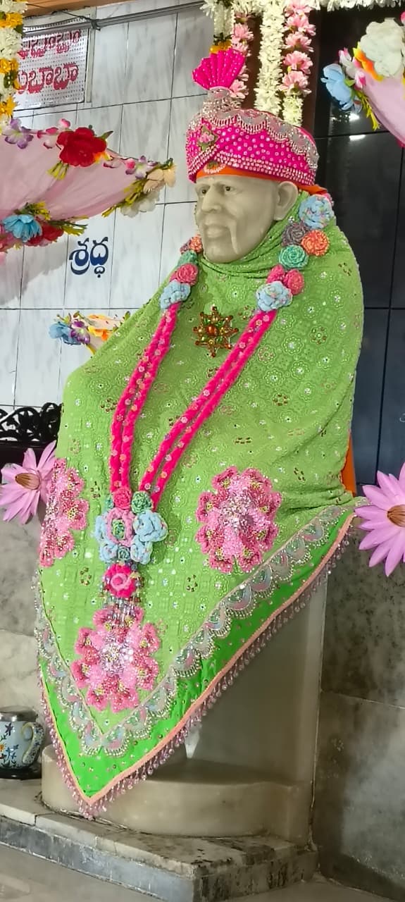 Closer view of the murti dressed in a green shawl with a pink beaded turban, the Shraddha signage visible on the wall