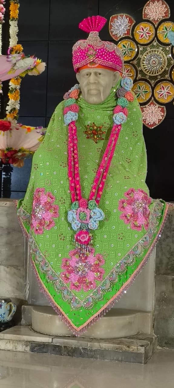The murti dressed in a green shawl with a pink pom-pom garland, a decorative round floral medallion visible on the wall behind