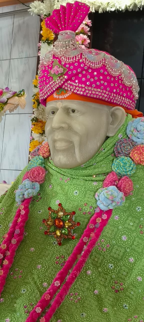 Close-up of the murti's face and pink jeweled turban with a fan-shaped crest, the green shawl with a crochet flower garland below