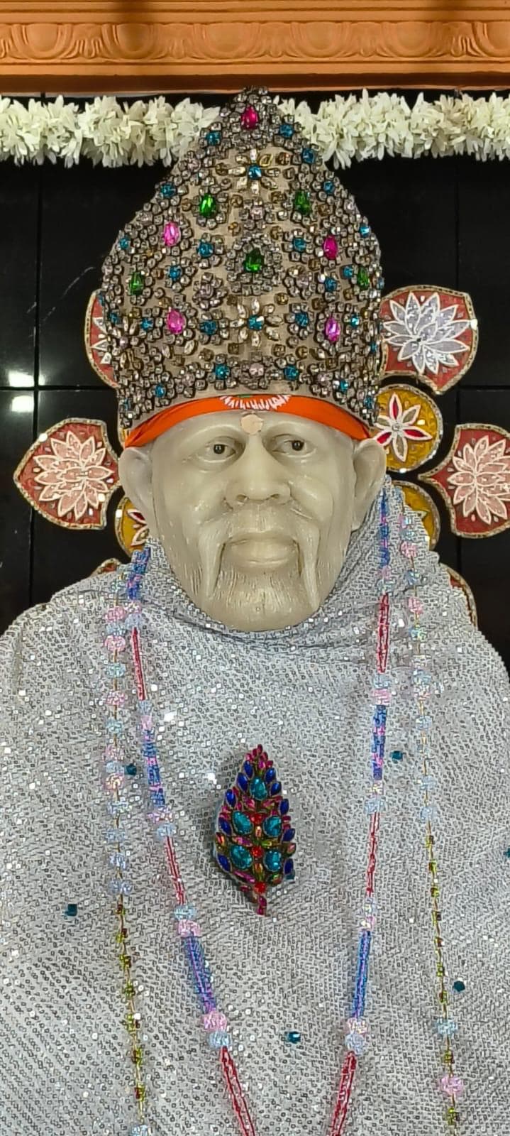 Close view of the murti's face and the same jeweled multicolor crown, now dressed in a silver sequined shawl, with pastel beaded garlands and a floral brooch, framed by decorative floral medallions on the wall behind