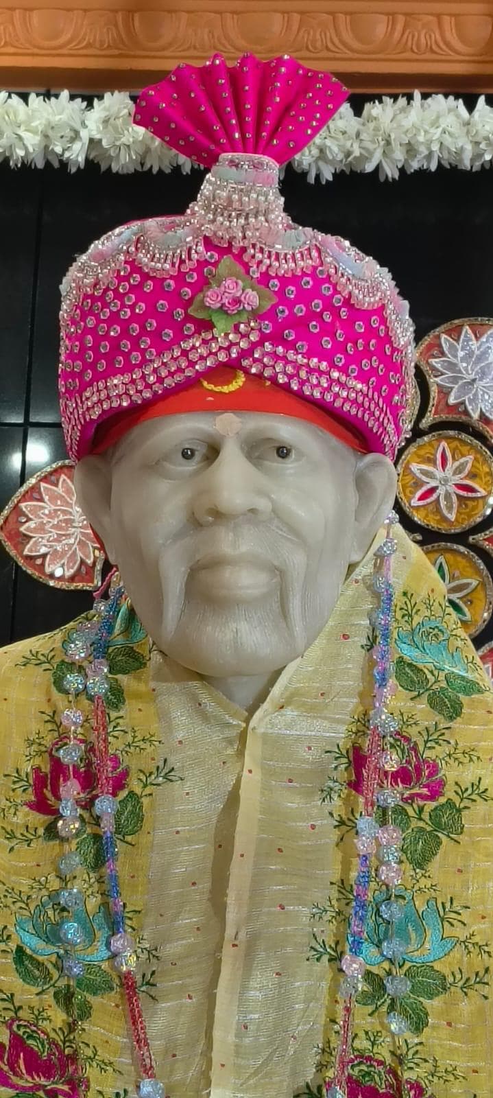 Close view of the murti's face and hot pink beaded turban with a fan-shaped crest, dressed in a pale yellow shawl with rose and teal floral embroidery