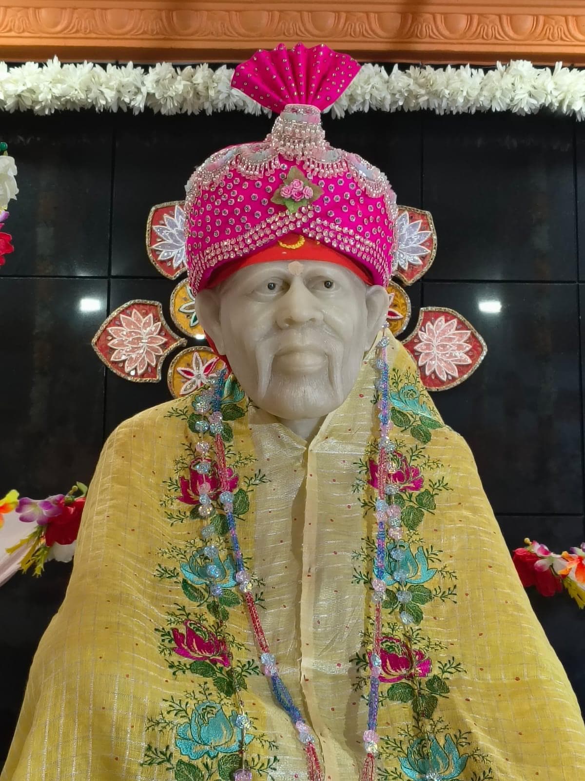 Another close view of the murti's face and hot pink beaded turban, dressed in the pale yellow floral-embroidered shawl, decorative floral medallions visible on the wall behind