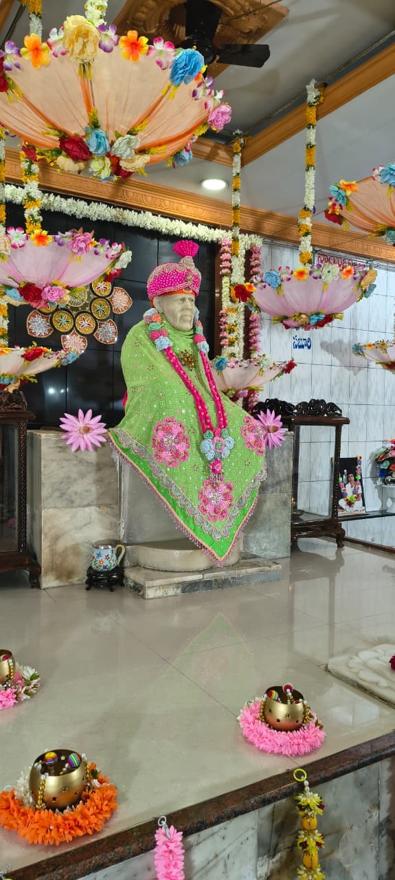 Wide view of the murti dressed in a green shawl with a pink beaded turban and pom-pom garland, hanging floral umbrellas above and small gold kalash pots decorated with flowers on the floor