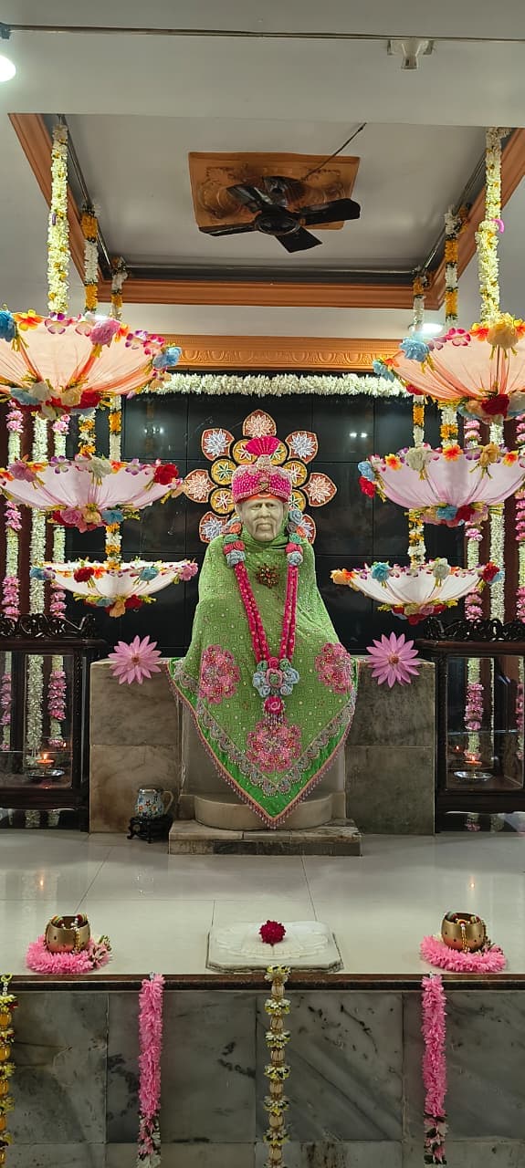Wide view of the sanctum for Guru Purnima, the murti dressed in a green shawl and pink beaded turban, framed by a decorative floral halo and hanging floral umbrellas