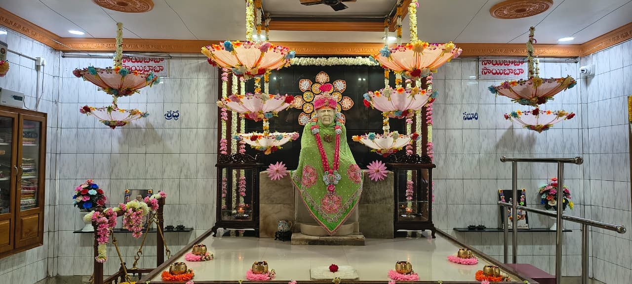 Wide view of the sanctum for Guru Purnima, the murti dressed in a green shawl with a pink pom-pom garland, a decorative floral halo behind, hanging floral umbrellas, and Shraddha and Saburi signage on the side walls