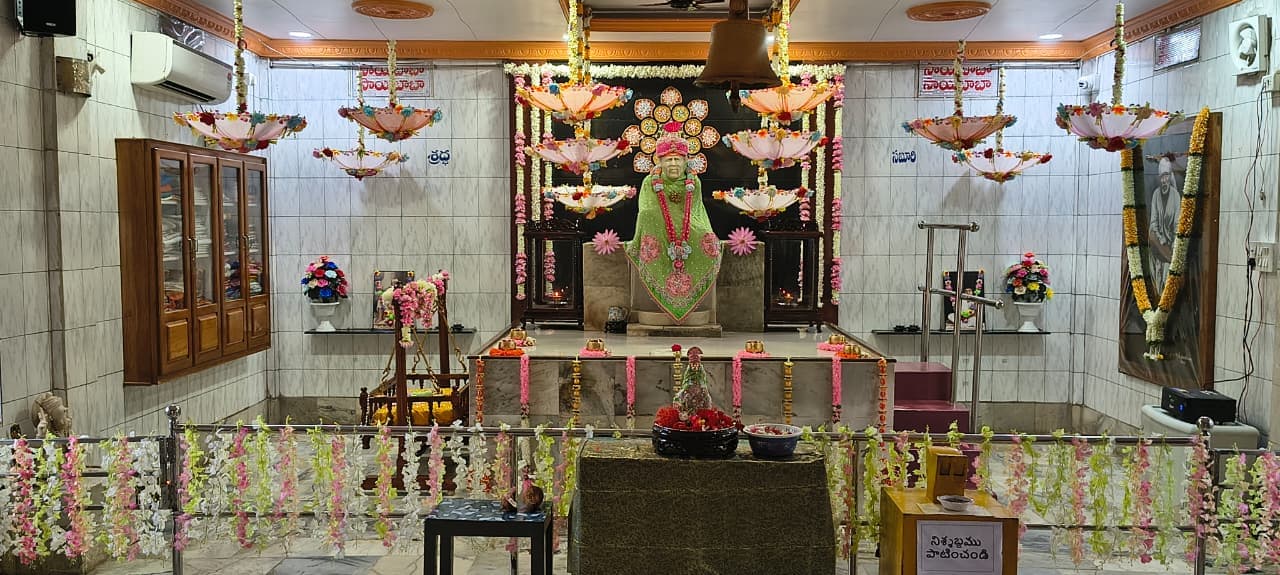 Wide view of the full sanctum for Guru Purnima, with a hanging bell above, a garland-decorated railing in the foreground, the small murti in a bowl of flowers on a pedestal, and the main murti dressed in a green shawl framed by a decorative floral halo and hanging umbrellas