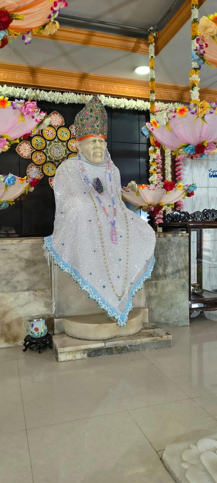 Angled wide view of the sanctum for Guru Purnima, the murti dressed in a silver sequined shawl with a light blue lace trim, pink garland strands hanging at the sides and colorful floral umbrellas visible nearby