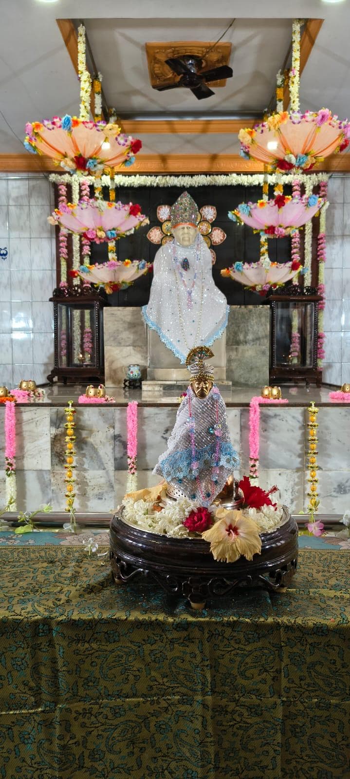 Wide view of the full sanctum for Guru Purnima, hanging floral umbrellas above, the main murti dressed in a silver sequined shawl in the background, and the small processional murti dressed in matching silver and blue lace on a round pedestal of flowers in the foreground