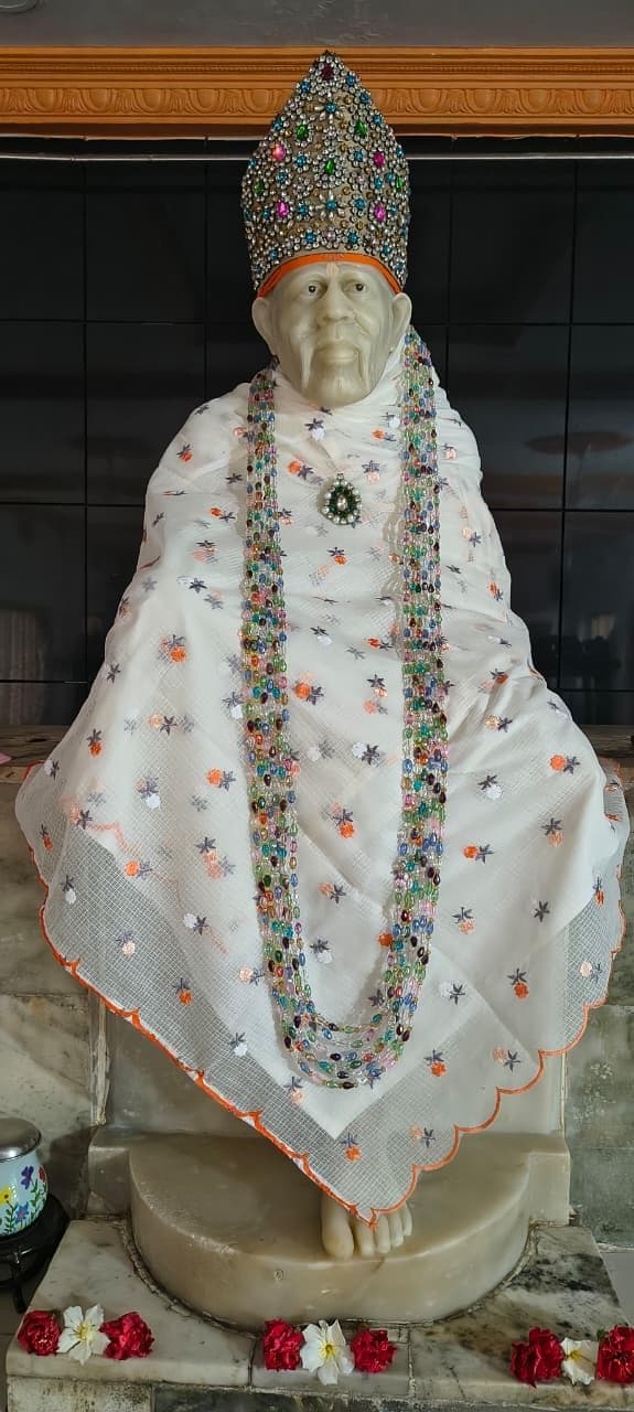 Wide view of the murti dressed in a sheer white shawl with an orange floral-star embroidery pattern and a scalloped orange-trimmed edge, wearing a heavily jeweled multicolor crown, with layered rainbow beaded garlands and fresh flowers placed at the base