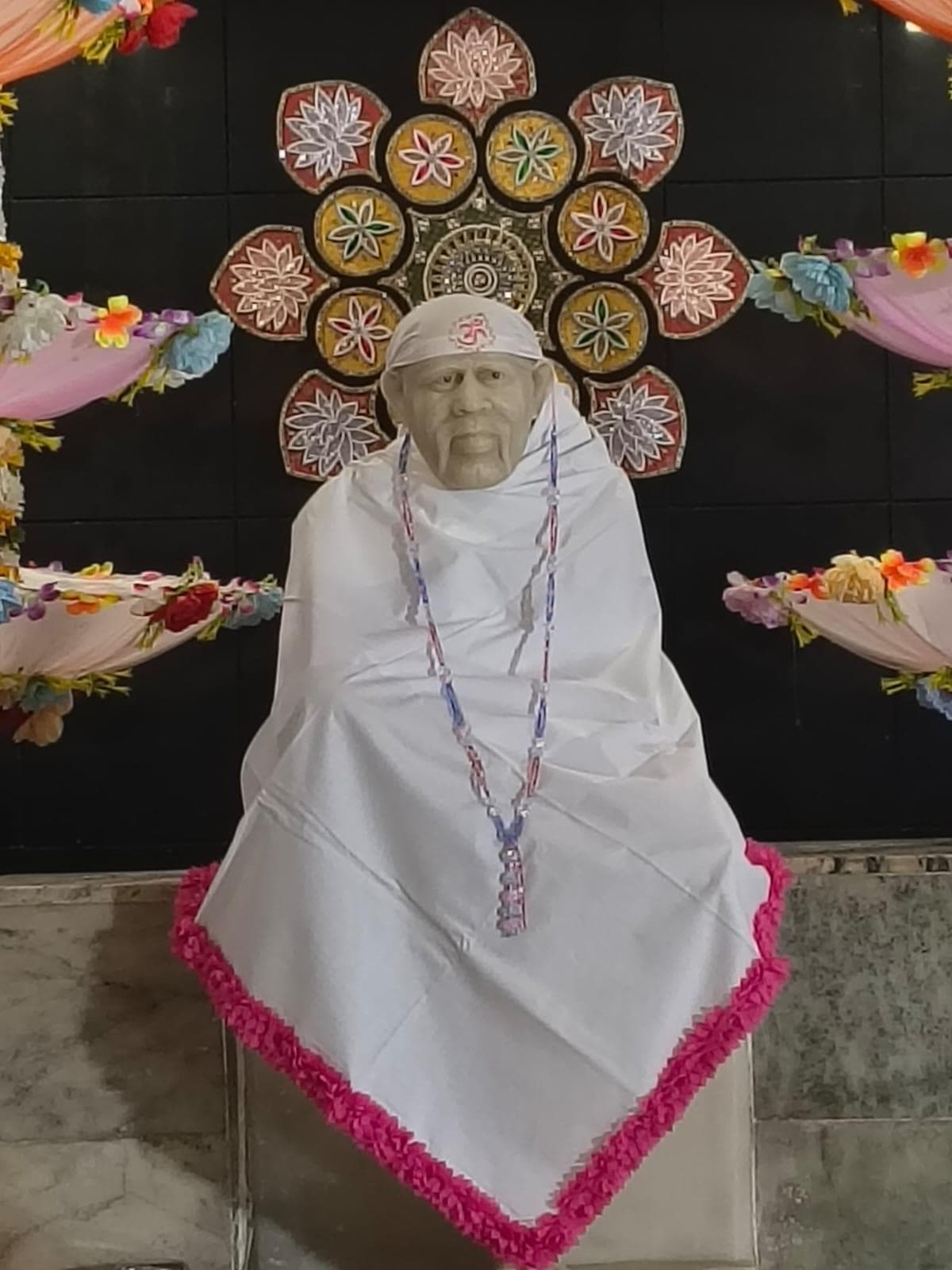 Wide view of the murti dressed in a plain white cloth with a hot pink ruffled trim and a beaded garland, framed by a large decorative round floral medallion on the wall behind