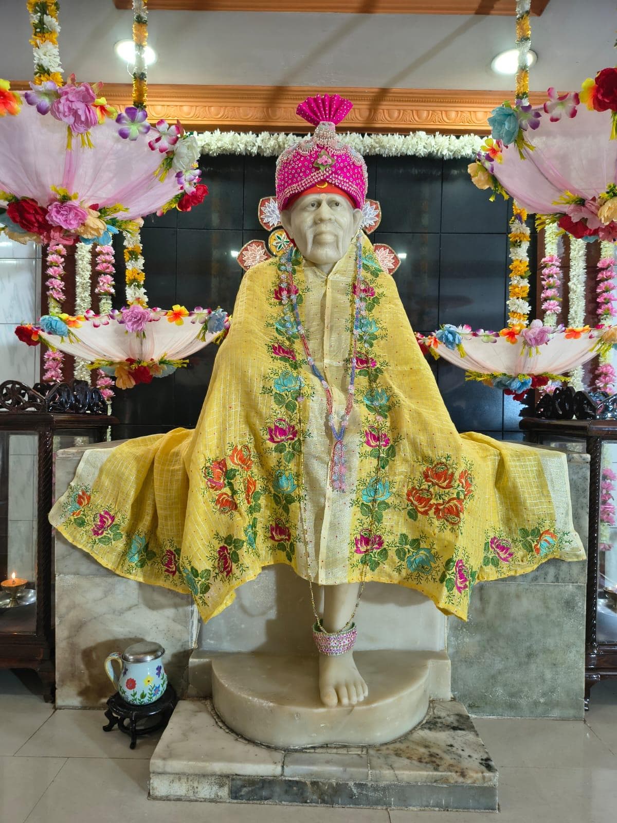 Wide view of the sanctum for Guru Purnima, the murti dressed in a pale yellow shawl with rose and teal floral embroidery and a hot pink beaded turban with a fan-shaped crest, hanging floral umbrellas above