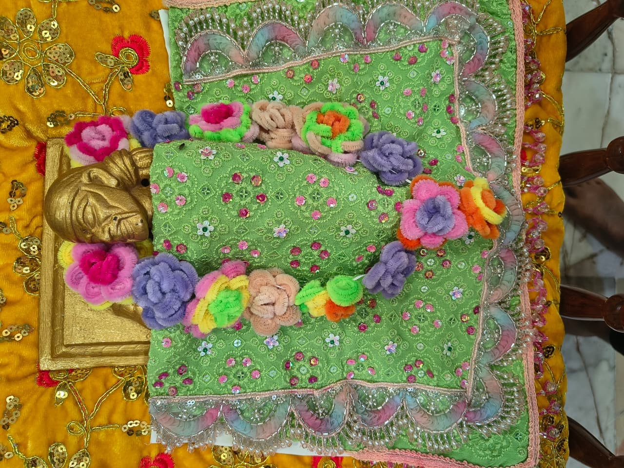A green sequined and beaded shawl with a colorful pom-pom flower garland laid out beside a small carved golden head, on a gold fabric backdrop, prepared ahead of dressing the murti for Guru Purnima