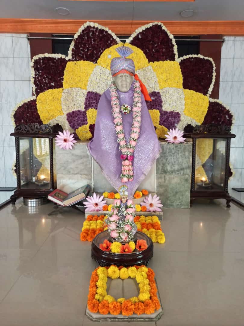 Wide view of the sanctum, the murti dressed in a lavender shawl and jeweled fan-crest turban, framed by a large floral peacock-shaped backdrop in maroon, yellow, and white, with a small utsav murti and marigold arrangement at the base