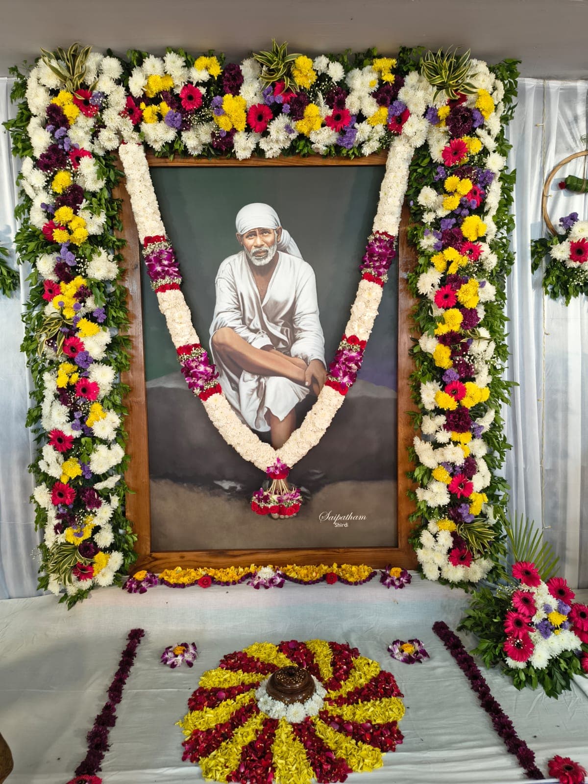 A framed painting of Sai Baba seated on a rock, signed "Saipatham Shirdi," bordered by a garland arch of white, yellow, and purple flowers, with a marigold and rose petal rangoli laid on the floor beneath it