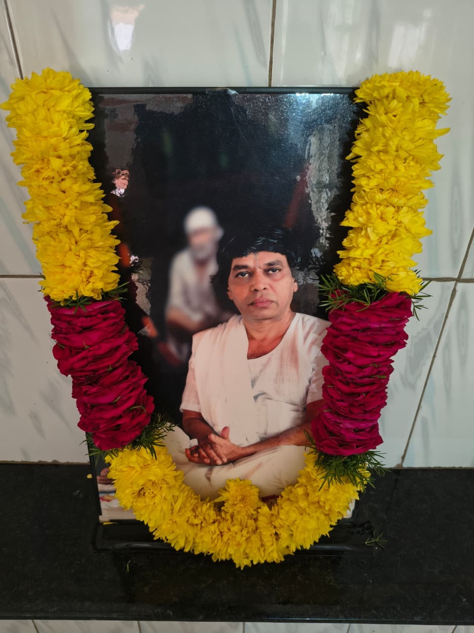 A framed photo of the guru figure seated in white, a blurred portrait of Sai Baba visible behind him, garlanded with yellow marigolds and pink roses