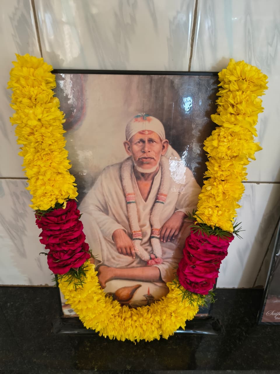 A framed traditional painted portrait of Sai Baba seated, garlanded with yellow marigolds and pink roses