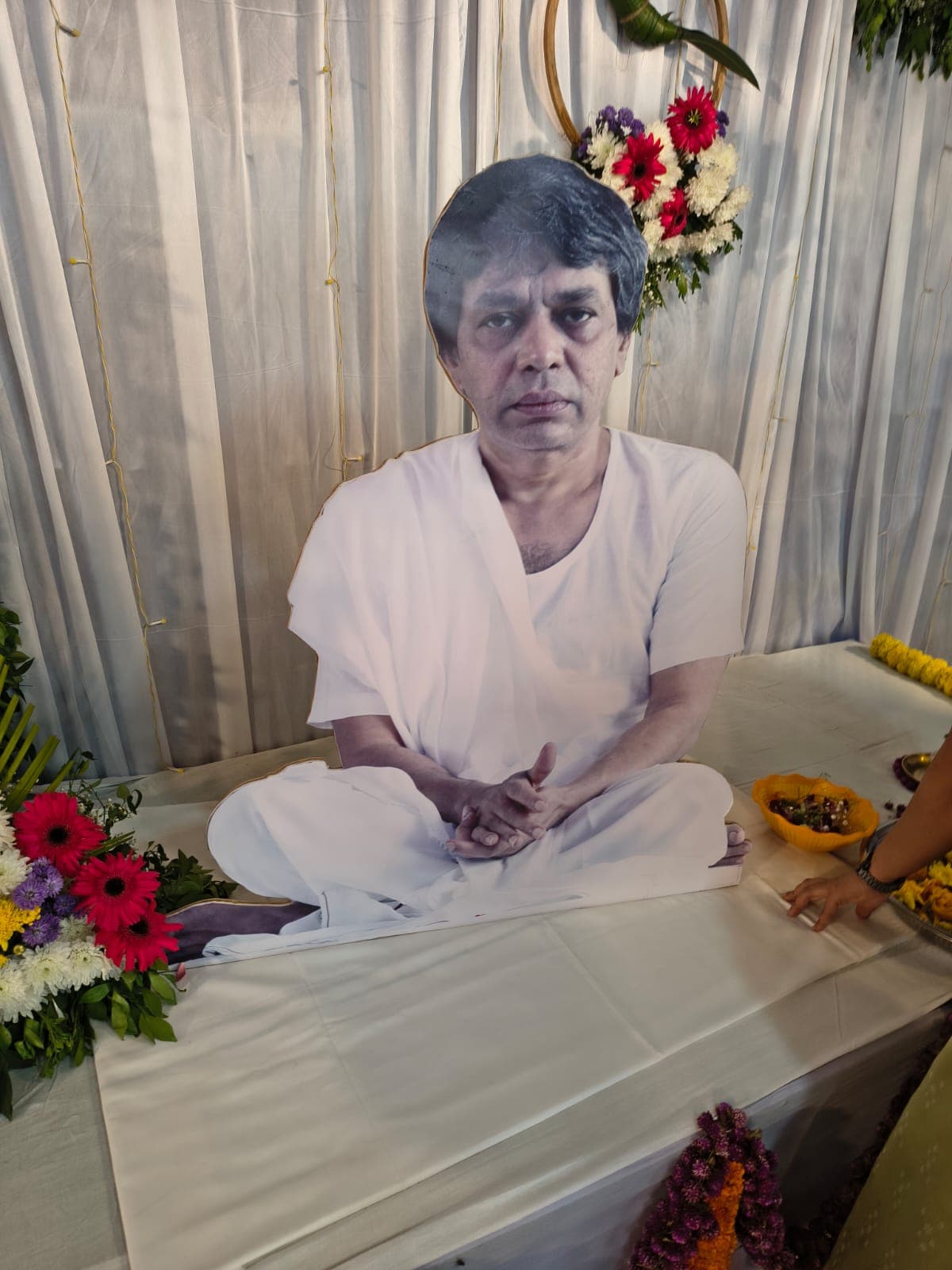 A life-size cutout photo standee of a seated figure in white, placed on a cloth-covered platform decorated with flower arrangements and a hanging ring wreath, in front of a curtained backdrop