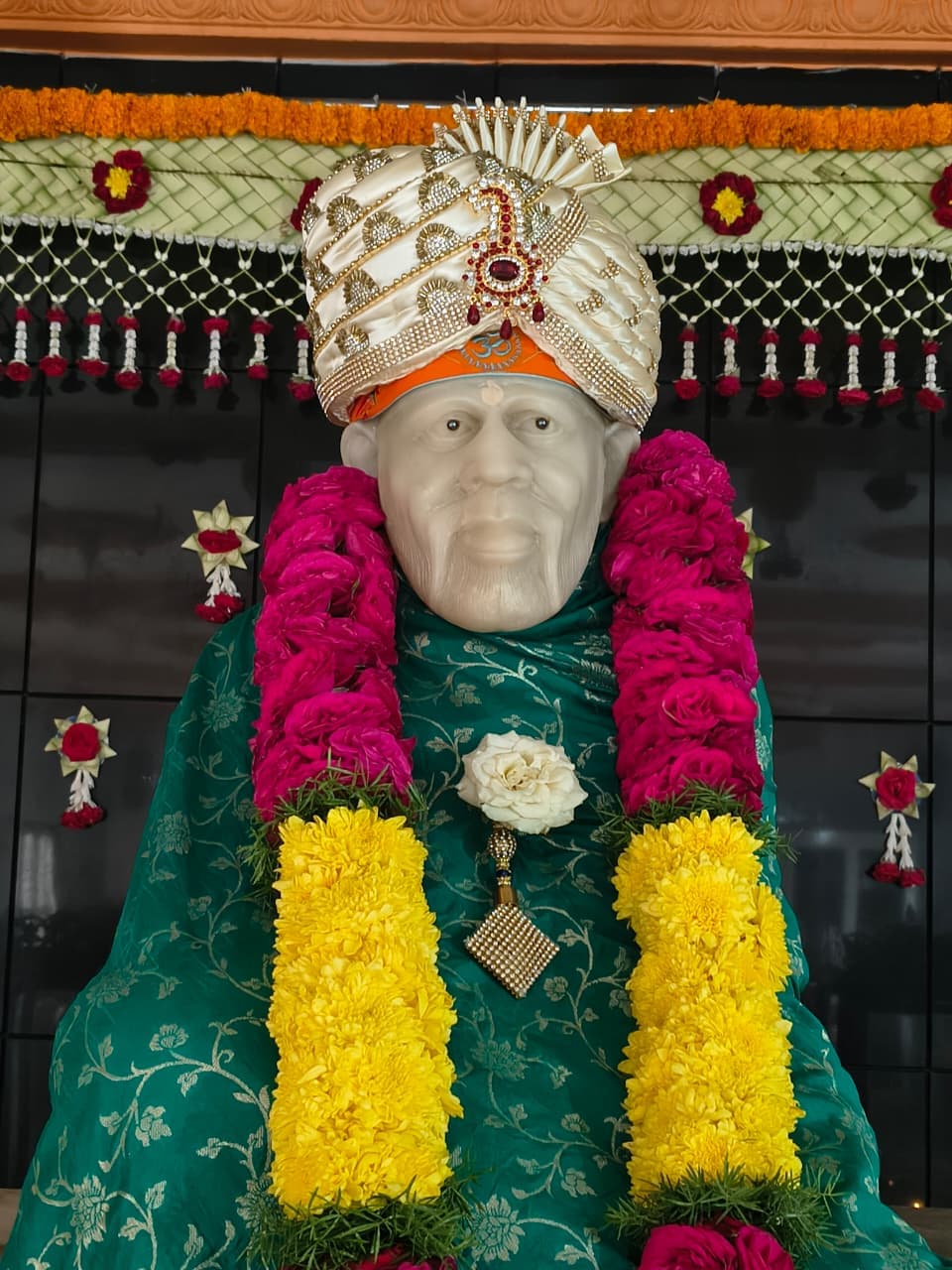 Close view of the murti's face and jeweled fan-crest turban with an "ॐ" mark, dressed in the green shawl with pink and yellow flower garlands